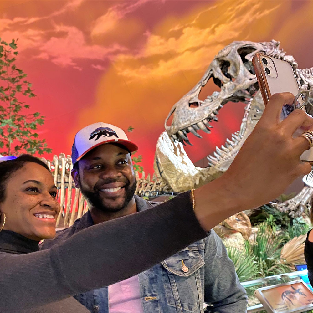 Two people posing for a selfie in front of Stan the T. rex in Dinosphere 